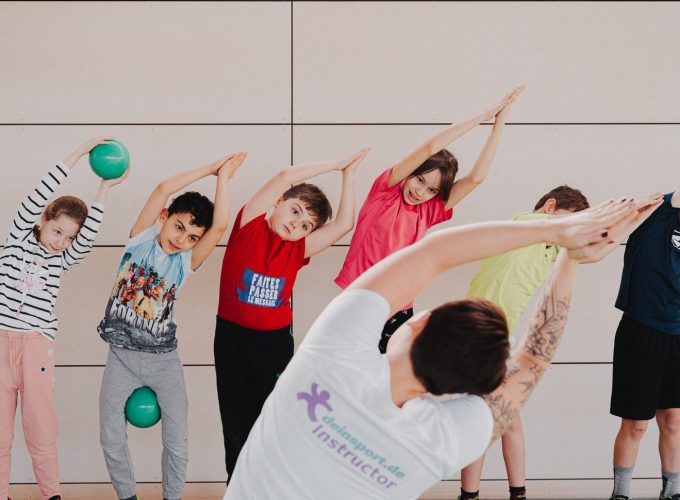 Sechs Kinder machen eine Bewegungsübung. Sie strecken ihre Arme nach oben und legen ihre Handflächen über ihren Köpfen zusammen. Sie werden von einer erwachsenen Person angeleitet.