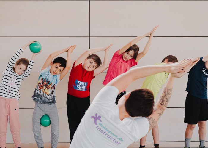 Sechs Kinder machen eine Bewegungsübung. Sie strecken ihre Arme nach oben und legen ihre Handflächen über ihren Köpfen zusammen. Sie werden von einer erwachsenen Person angeleitet.